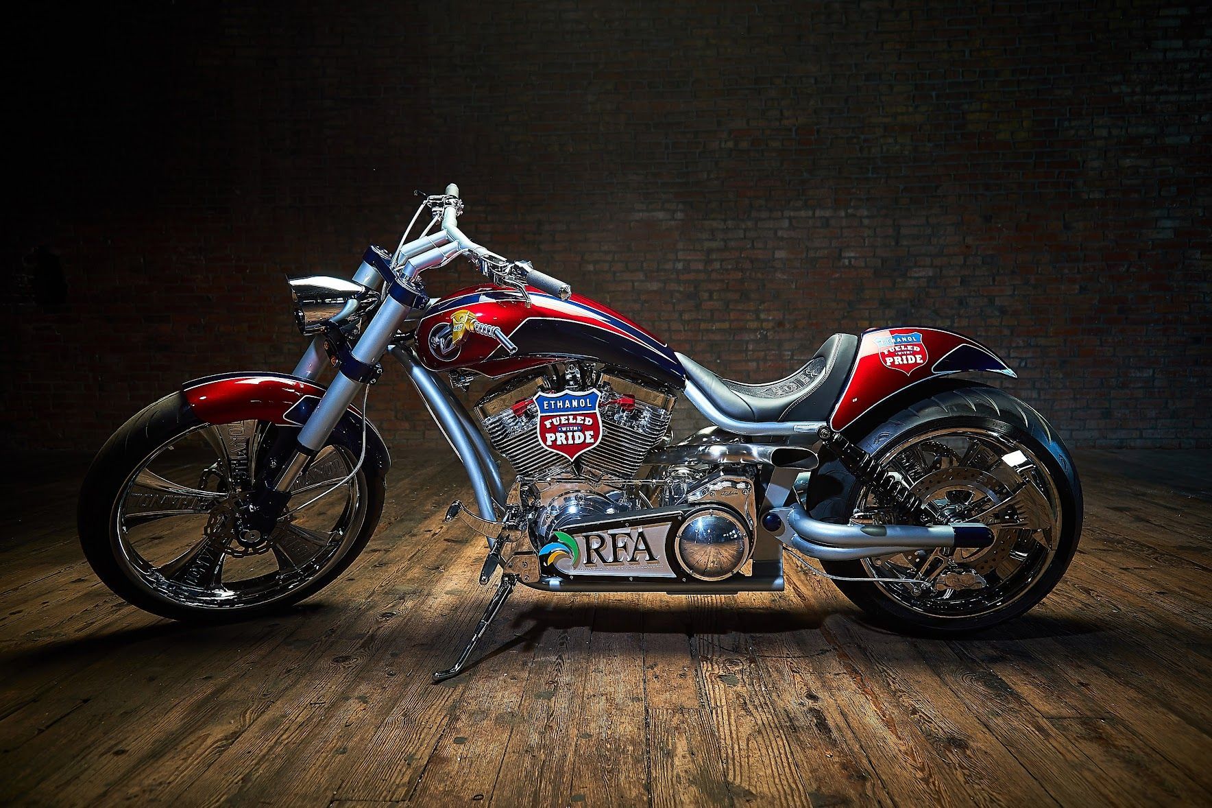 A red and black motorcycle is parked on a wooden floor in front of a brick wall.