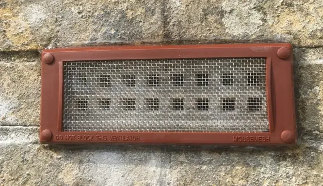 Brown rectangular vent cover with metal mesh screen on a stone wall.
