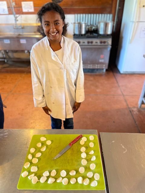 A Montessori child in a chef 's coat is standing in front of a cutting board