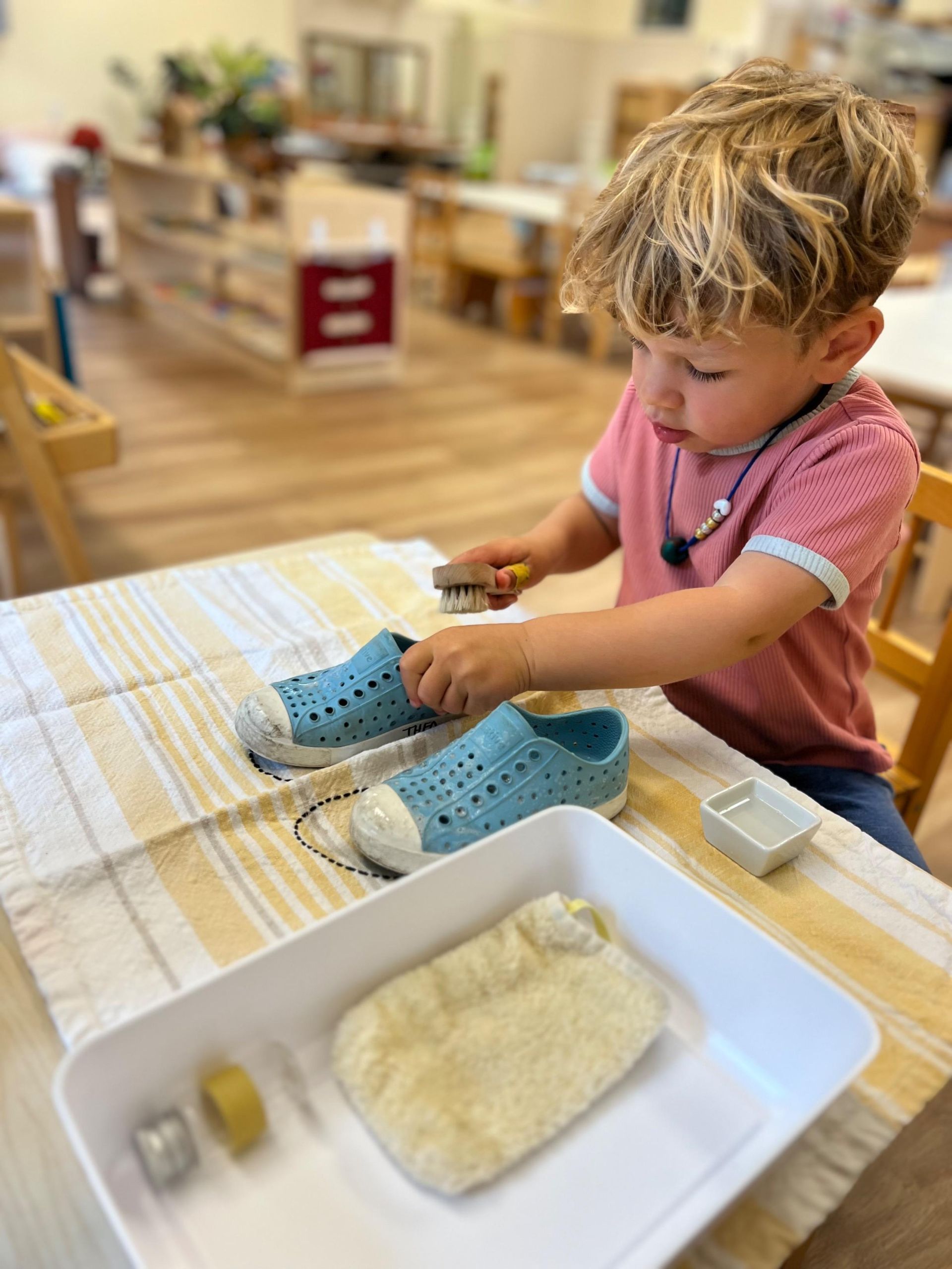 A Montessori child is cleaning a pair of shoes with a brush.