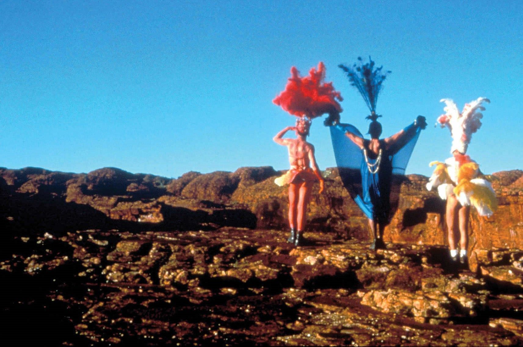 A trio of drag queens standing on a rocky hillside with feathers on their heads