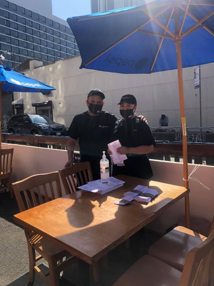 Two men wearing masks are standing next to a table under an umbrella.