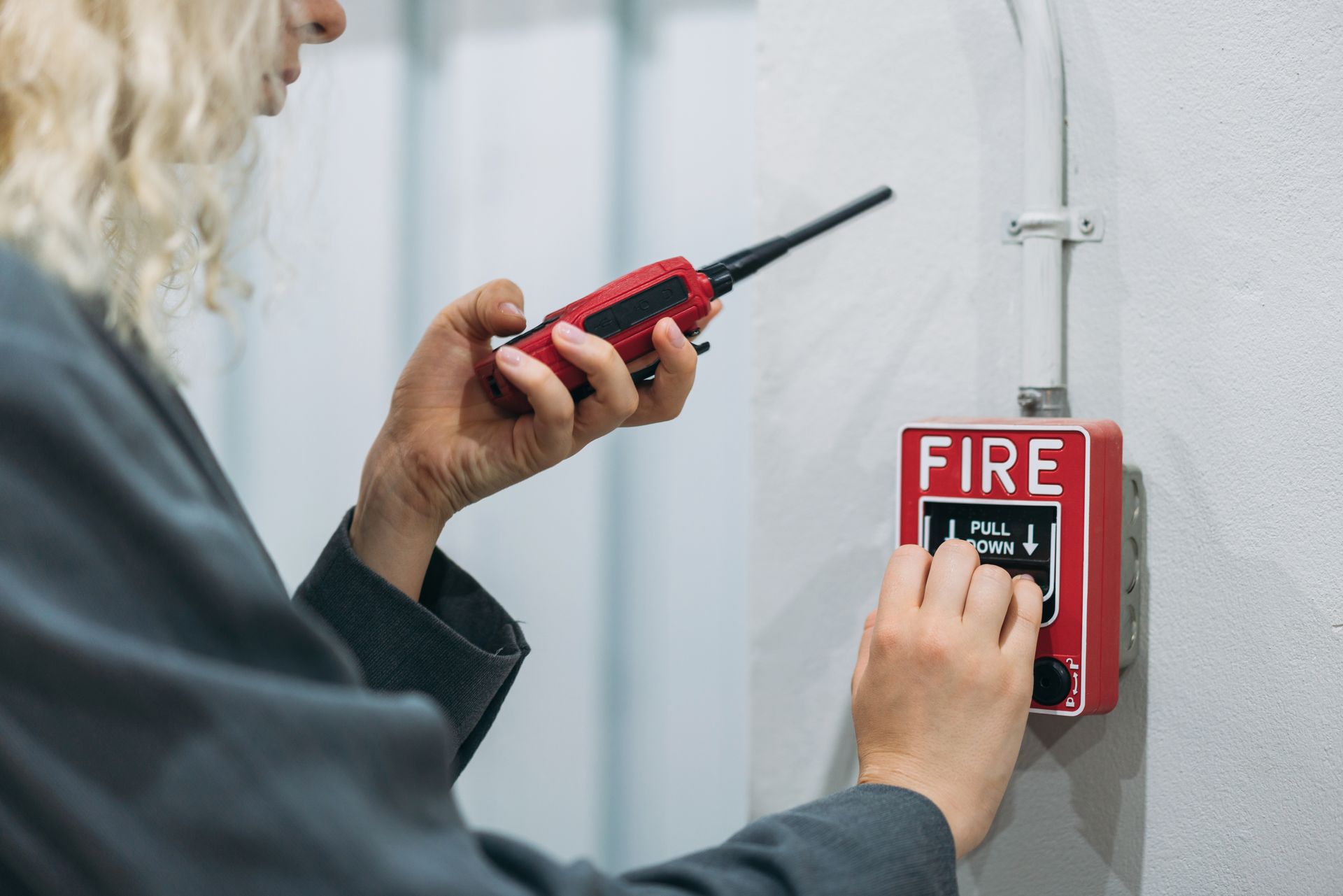 A woman is pressing a fire alarm button while holding a walkie talkie.