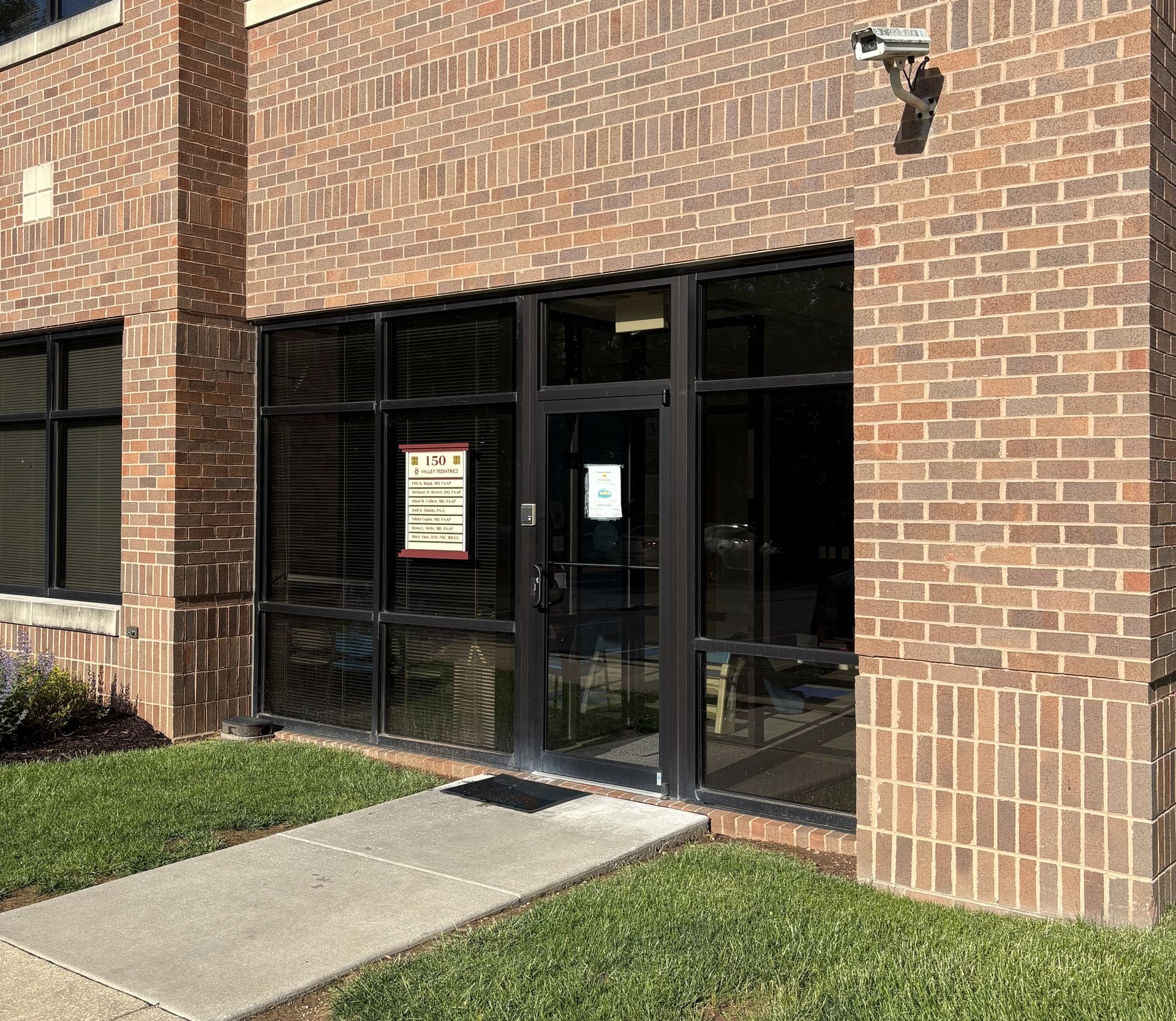Brick building entrance with black-framed glass doors and windows.  Concrete pathway and green grass.