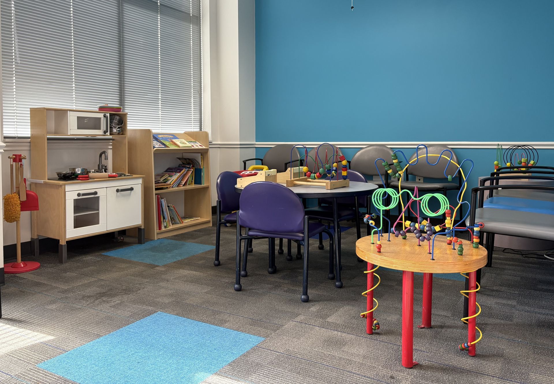 A play room with a toy kitchen, table, chairs, and a blue wall, carpet and blinds.