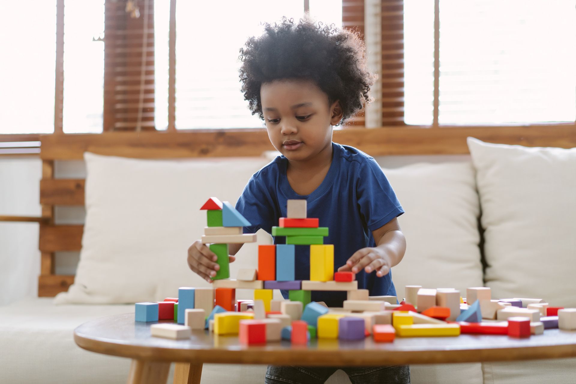 Young child with curly hair building a colorful tower with wooden blocks on a table.
