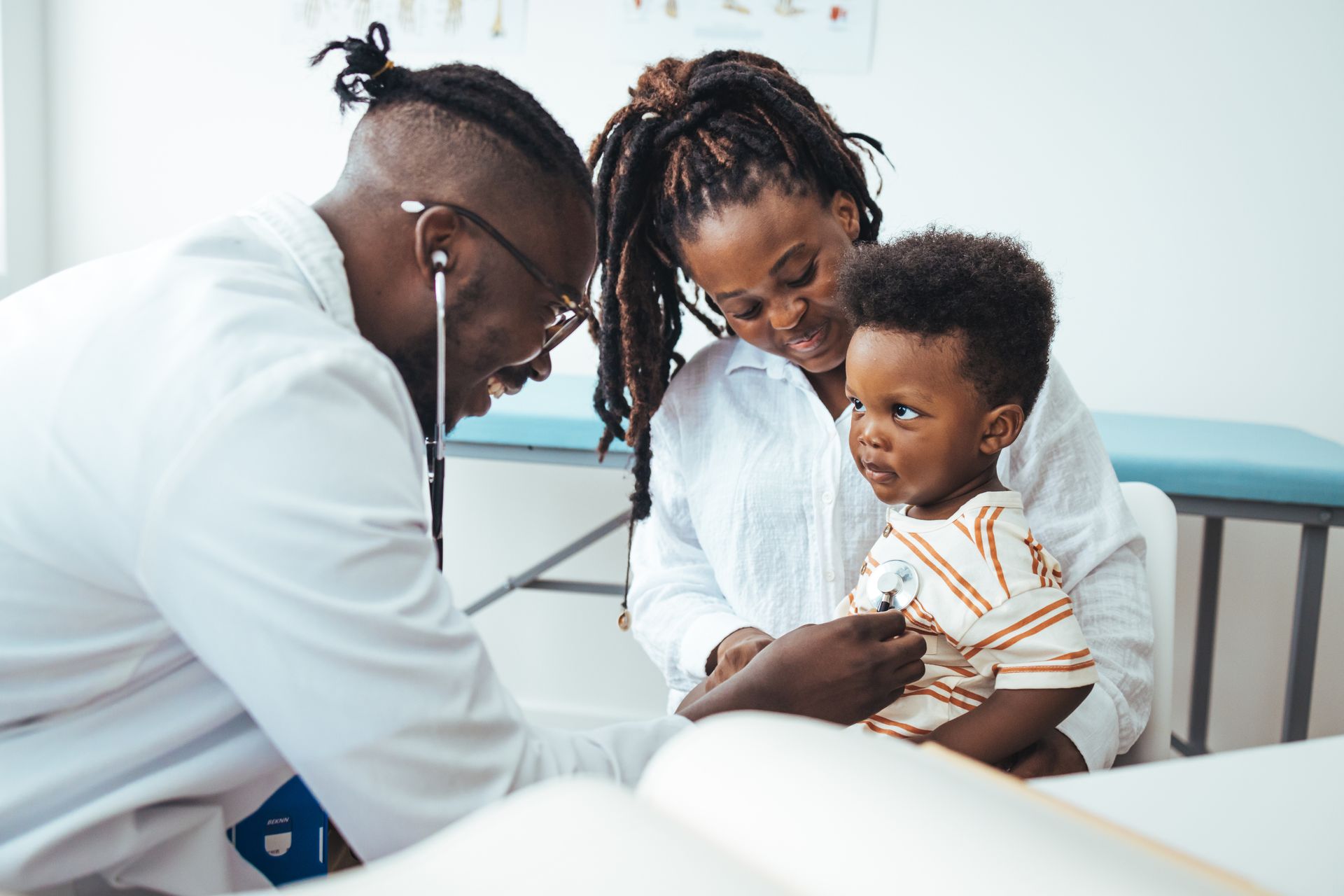 Black doctor using stethoscope on a child held by Black mother in a medical office.