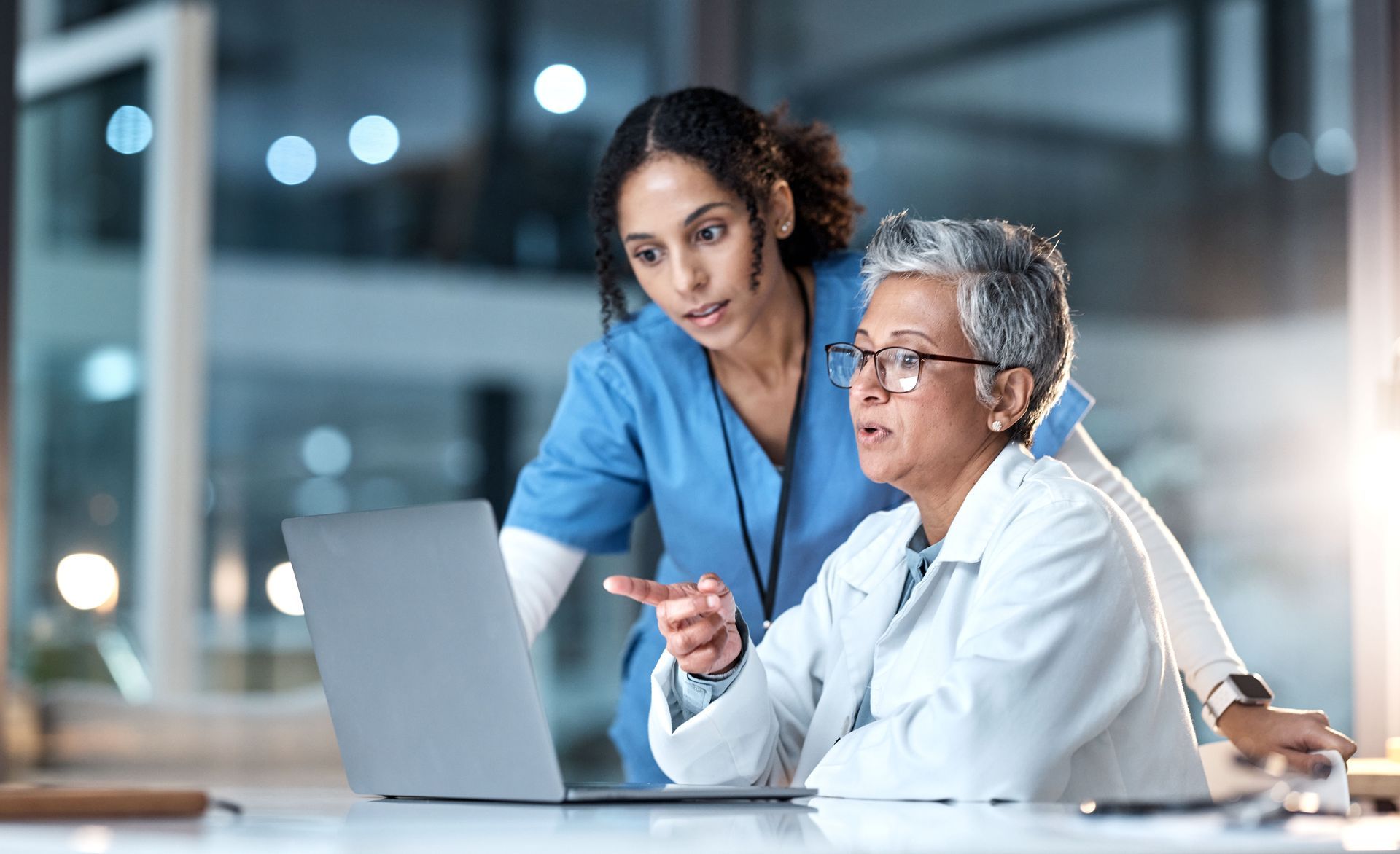 Two healthcare professionals, one young, one older, looking at laptop screen, pointing; lit interior.
