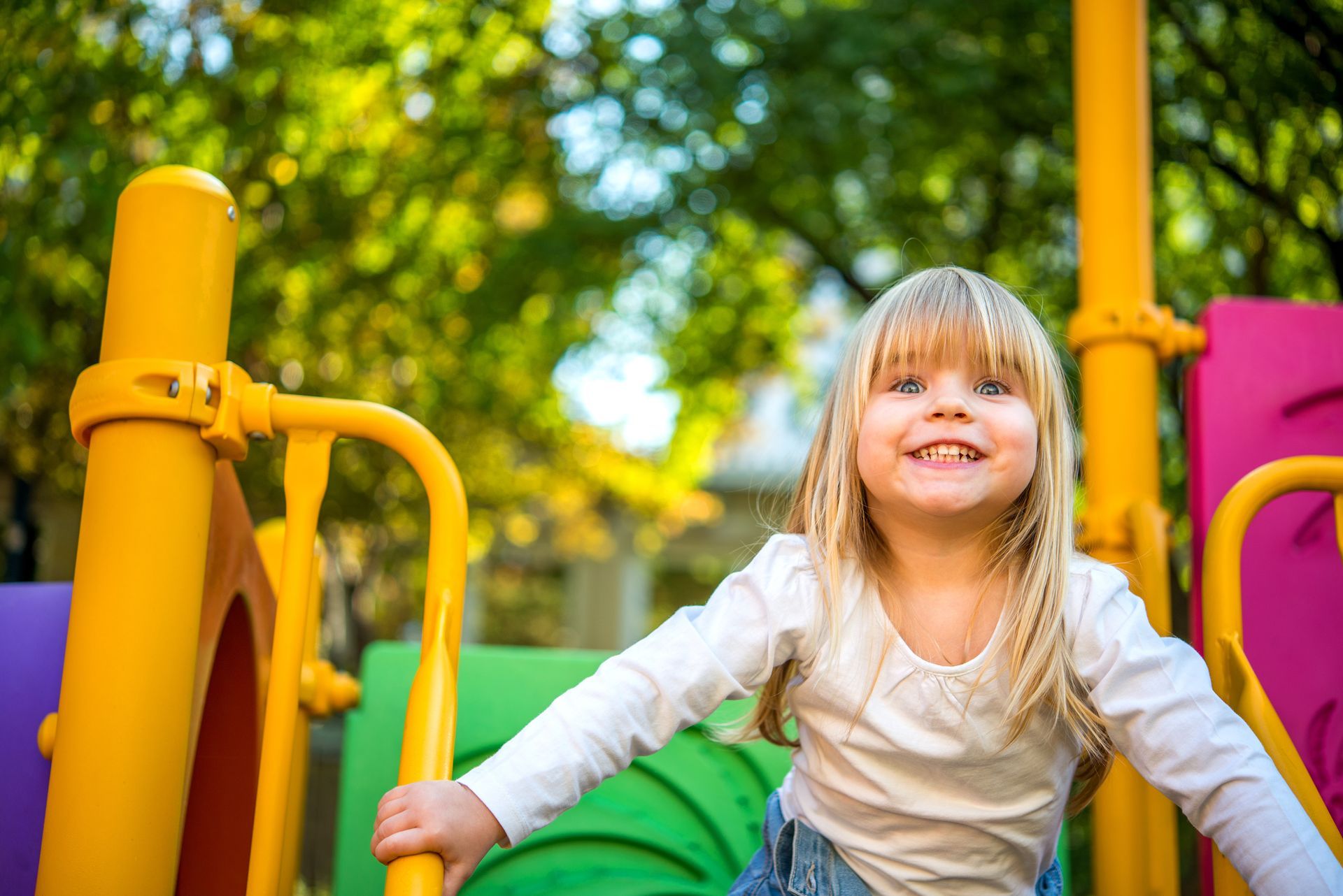 Blonde girl smiles, holding yellow bars on a colorful playground, green trees in the background.