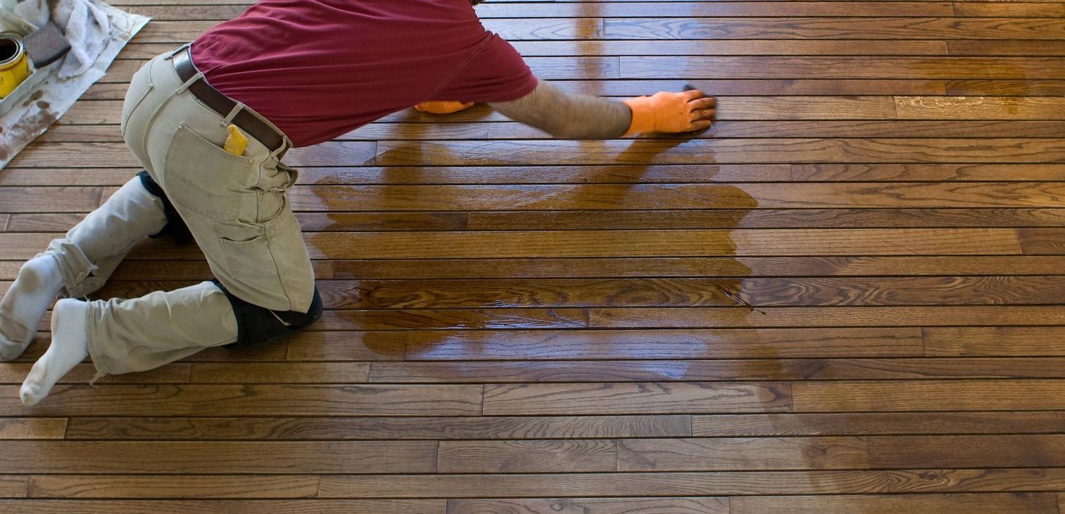 Person applying finish to a wood floor, kneeling. They wear a red shirt, orange gloves, and light pants.