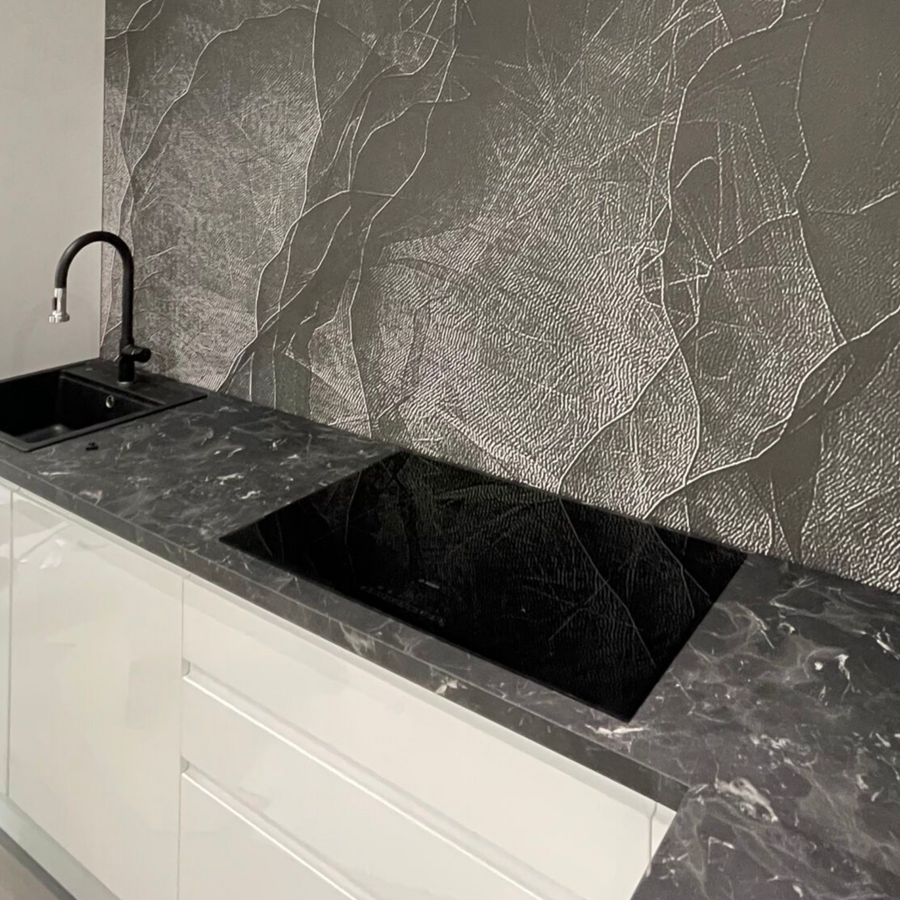 Cucina moderna con ripiani neri, piano cottura, lavello e paraschizzi metallico. Mobili bianchi.