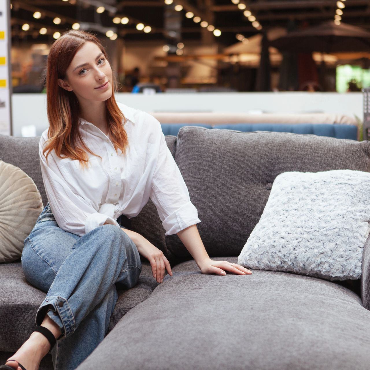 Donna sorridente in camicia bianca e jeans seduta su un divano grigio in un negozio di mobili.