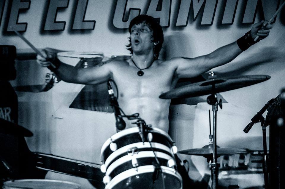 A shirtless man is playing drums in front of a sign that says el camilly