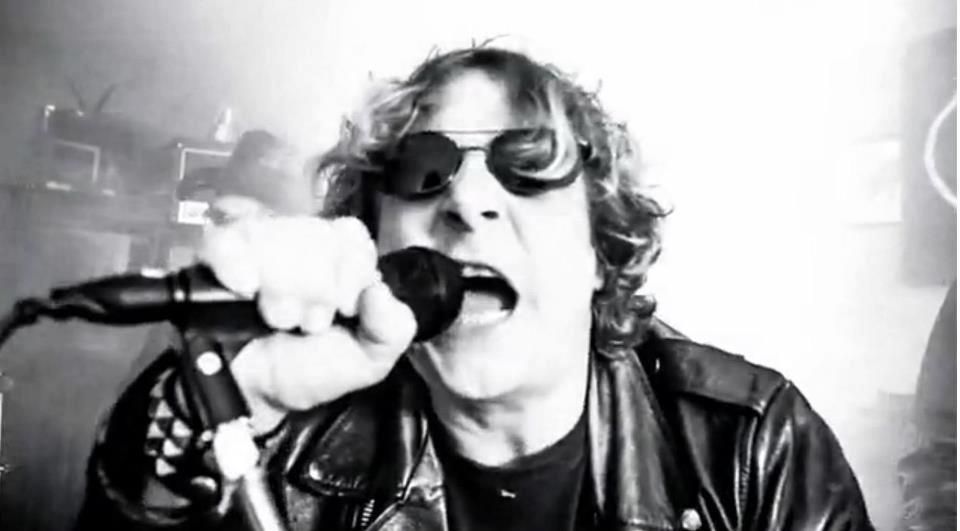 A man wearing sunglasses and a leather jacket is singing into a microphone.