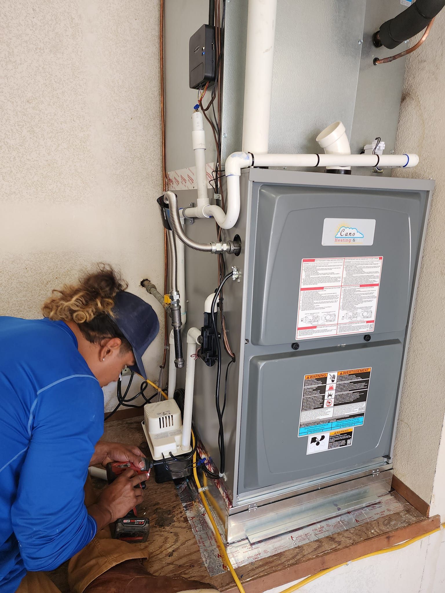 HVAC technician working on a furnace in a utility room; white pipes and gray furnace.