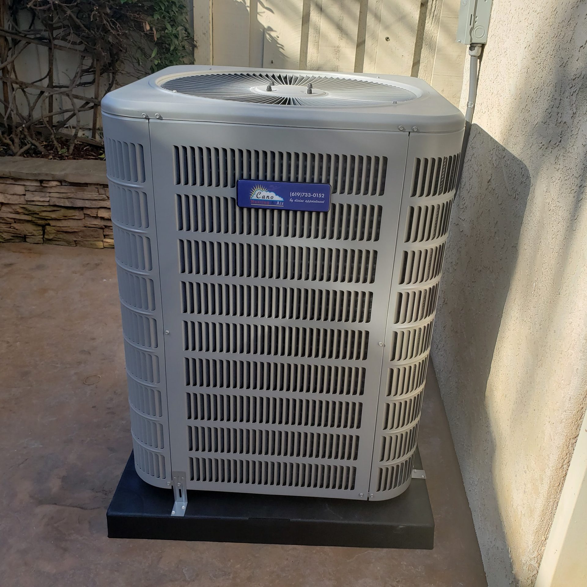 Gray air conditioning unit on a black platform against a beige wall and concrete ground.