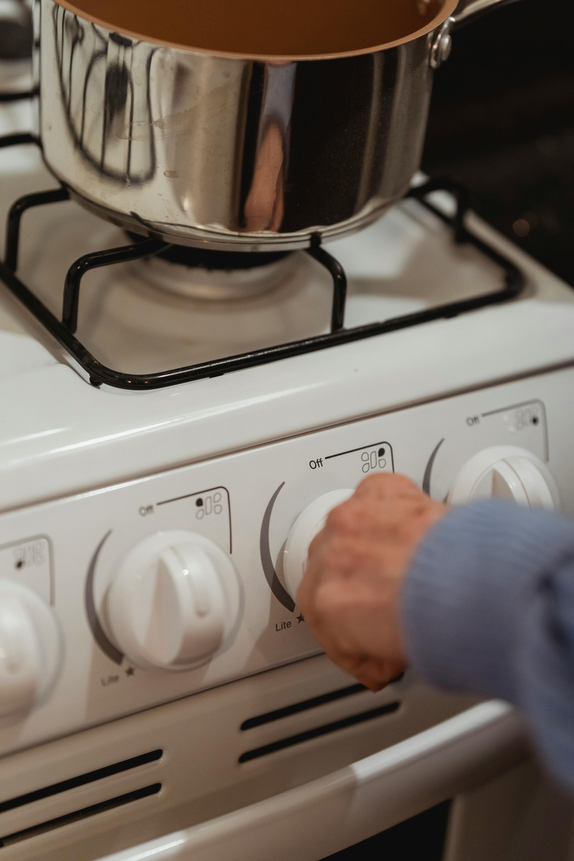 A hand turns a stove knob, with a pot on the burner; white stove, bright light.