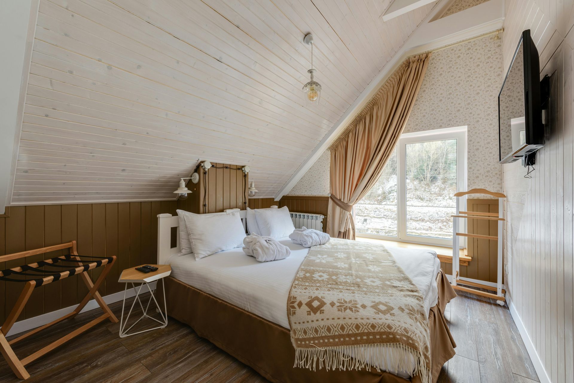 Bedroom with white slanted ceiling, bed with brown blanket, window with curtains, TV, luggage rack.