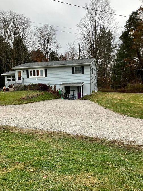 White split-level house with a gravel driveway, front lawn, and trees in the yard