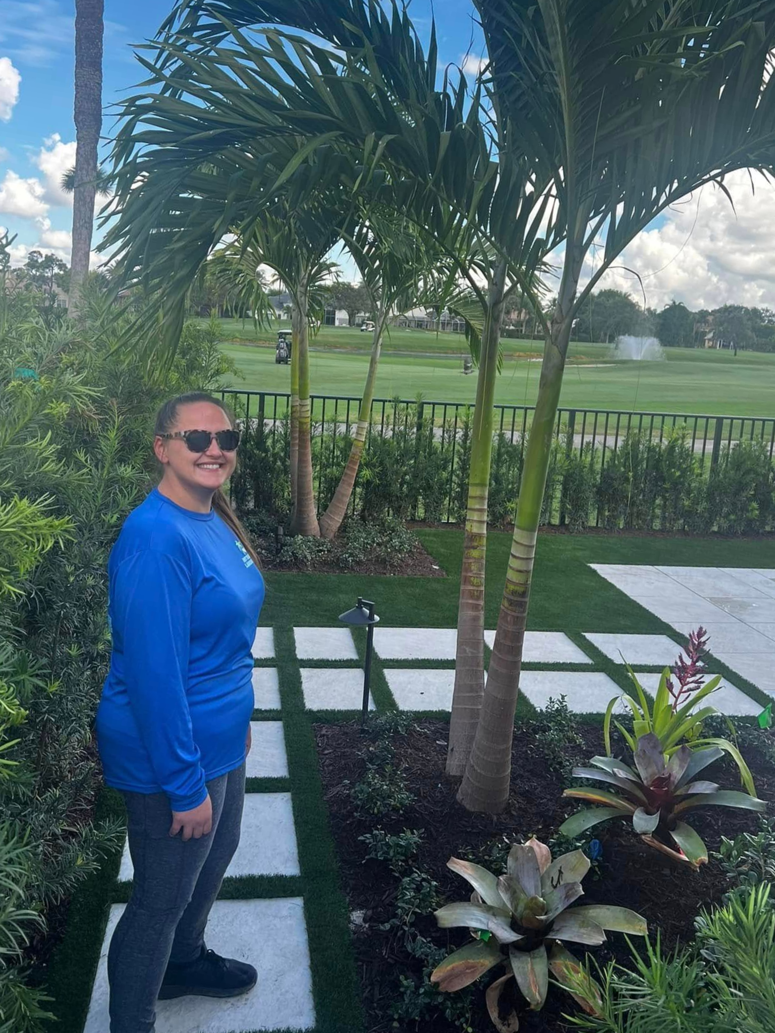 Person standing in a landscaped garden beside a palm tree and white stepping stones, smiling at the camera.
