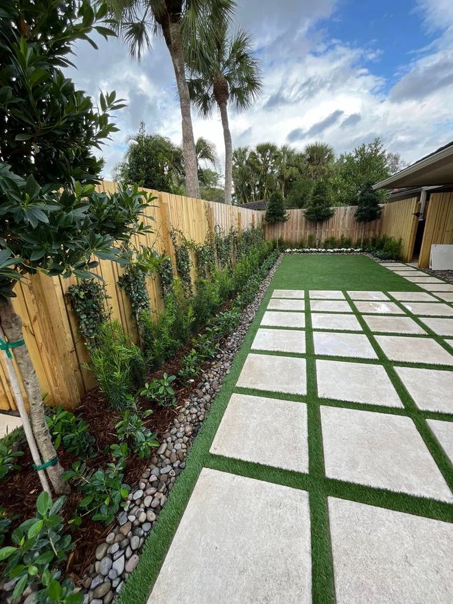 Modern landscaped backyard with white stepping stones, green grass grid, palm trees, and a wooden fence
