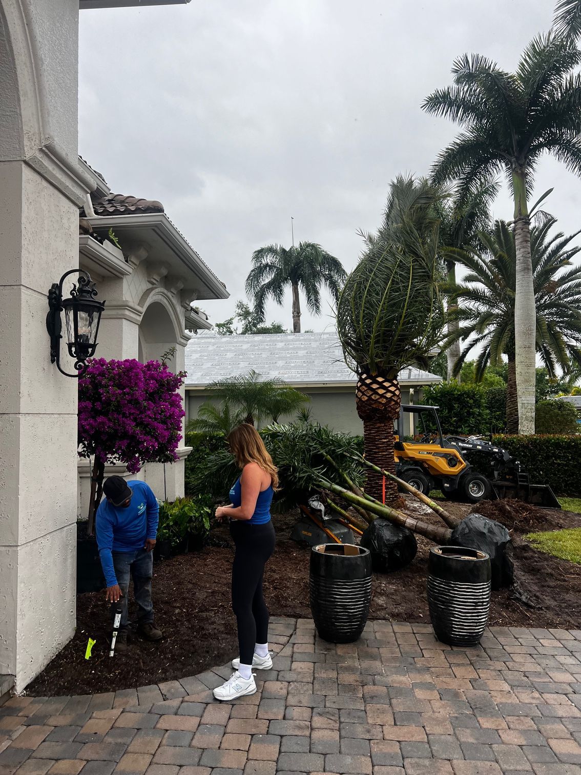 Two people landscaping near a home. A worker plants, and another watches. Potted plants and a lawnmower are nearby.