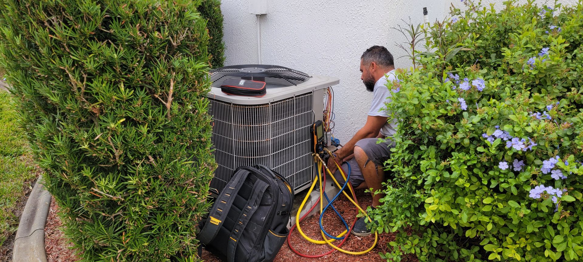 A person is working on an air conditioning unit outside a building, surrounded by greenery.