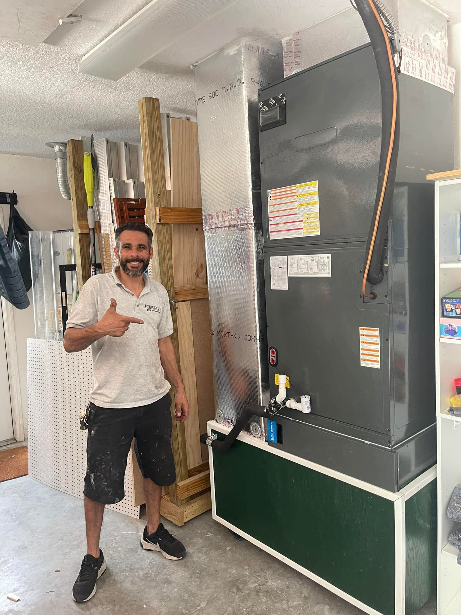 Man points to a new HVAC unit in a garage setting. Gray unit, silver ductwork, wood storage.