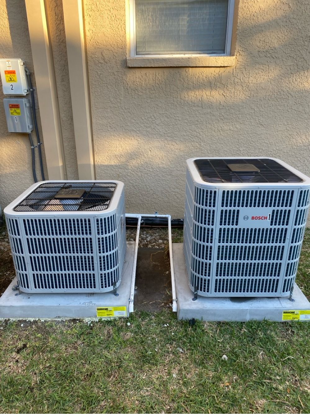 Two air conditioning units on concrete pads next to a building with a window and electrical box.