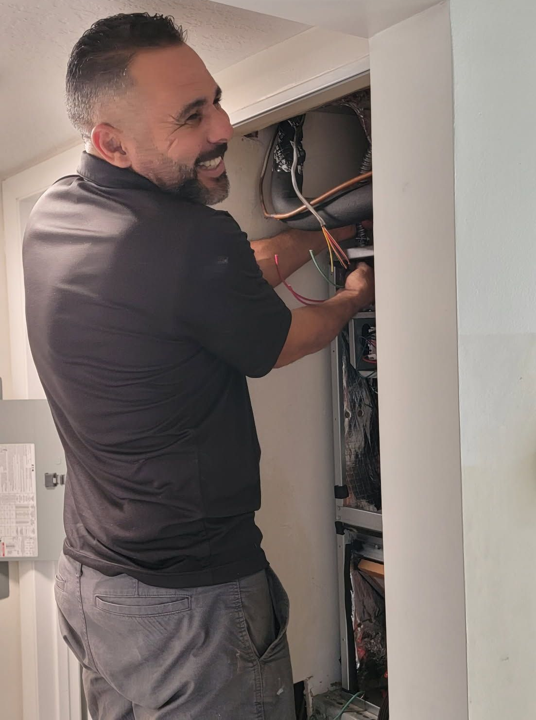 A man in a black shirt works on electrical wiring inside a wall cabinet, smiling.