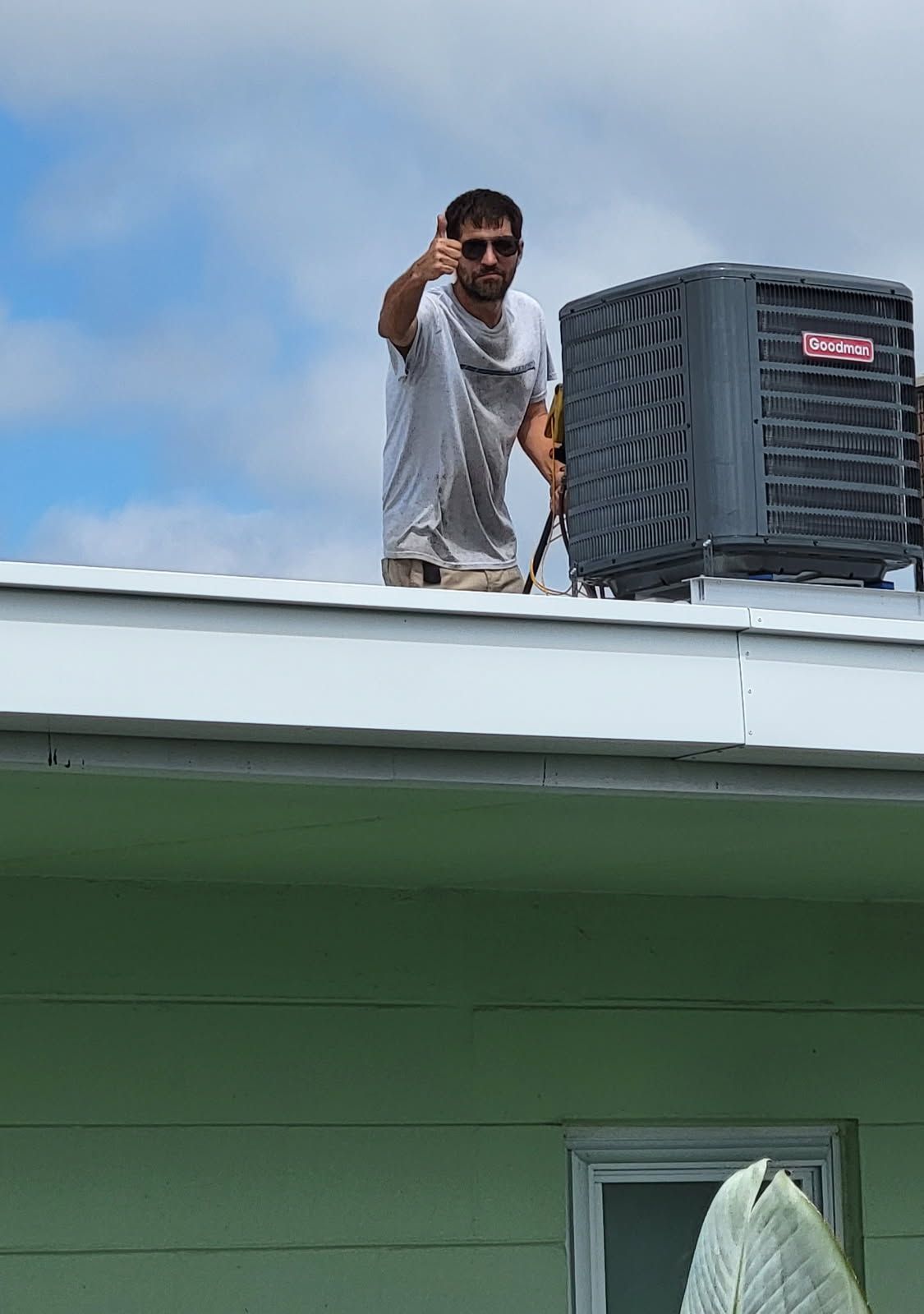 Man on a roof gives thumbs up next to an AC unit, against a partly cloudy sky.