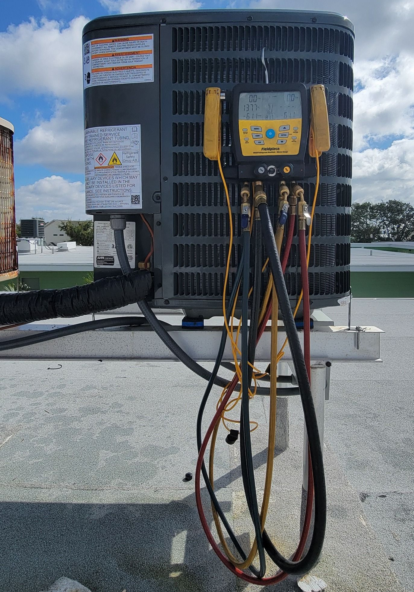 HVAC unit on a rooftop, with gauge manifold attached by multiple hoses, under a partly cloudy sky.