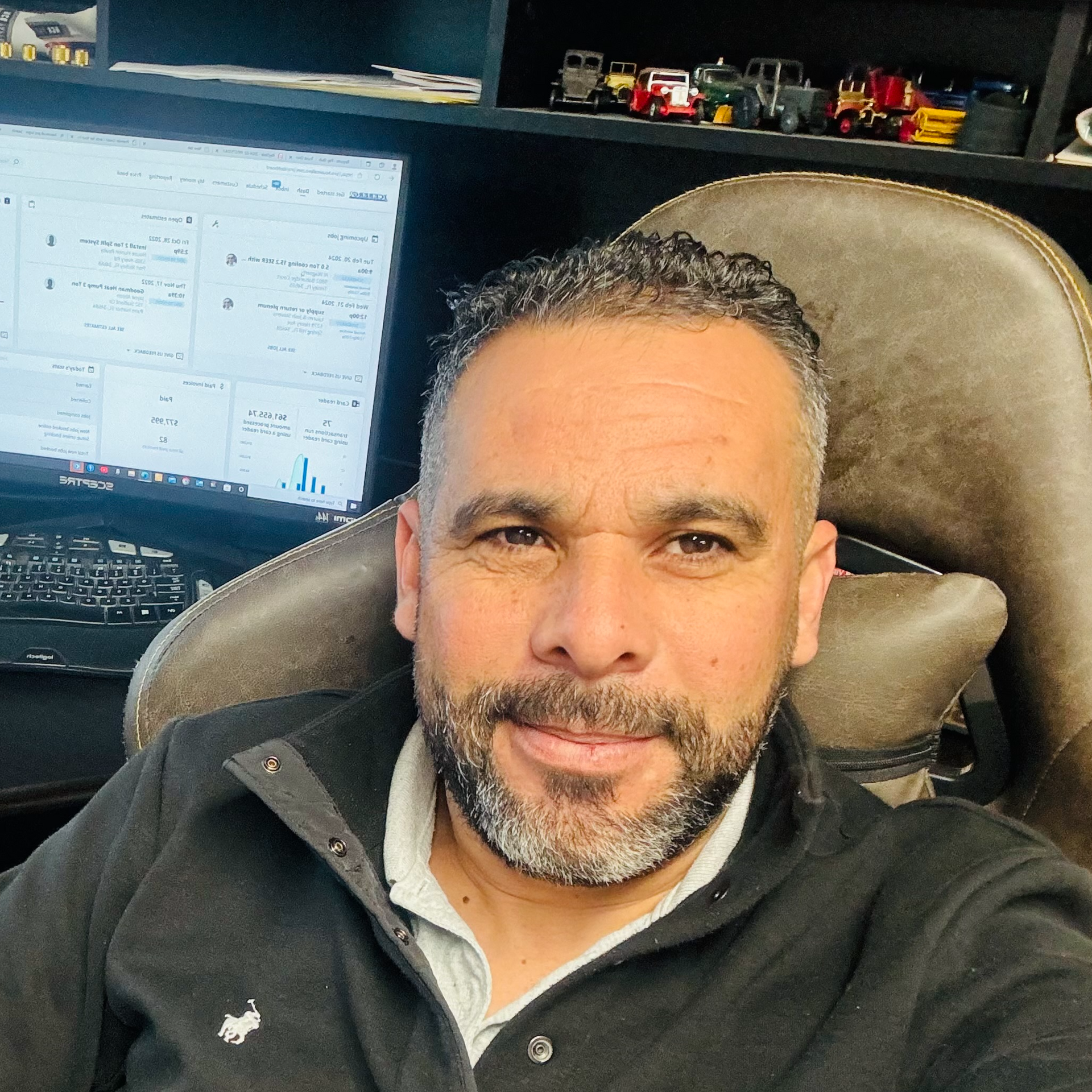 Man with a trimmed beard in a dark jacket sits in an office chair, working at a computer.