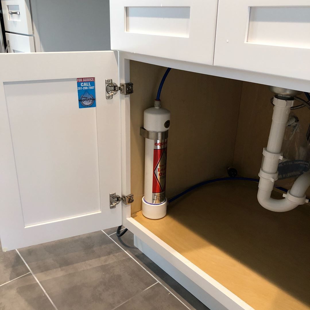White cabinet open, revealing a cylindrical water filter connected to plumbing under a sink.