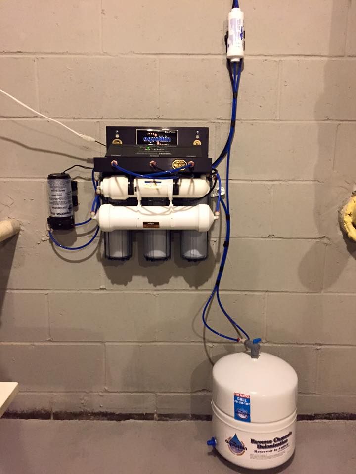 Water filtration system mounted on a gray cinder block wall with a storage tank on the floor.