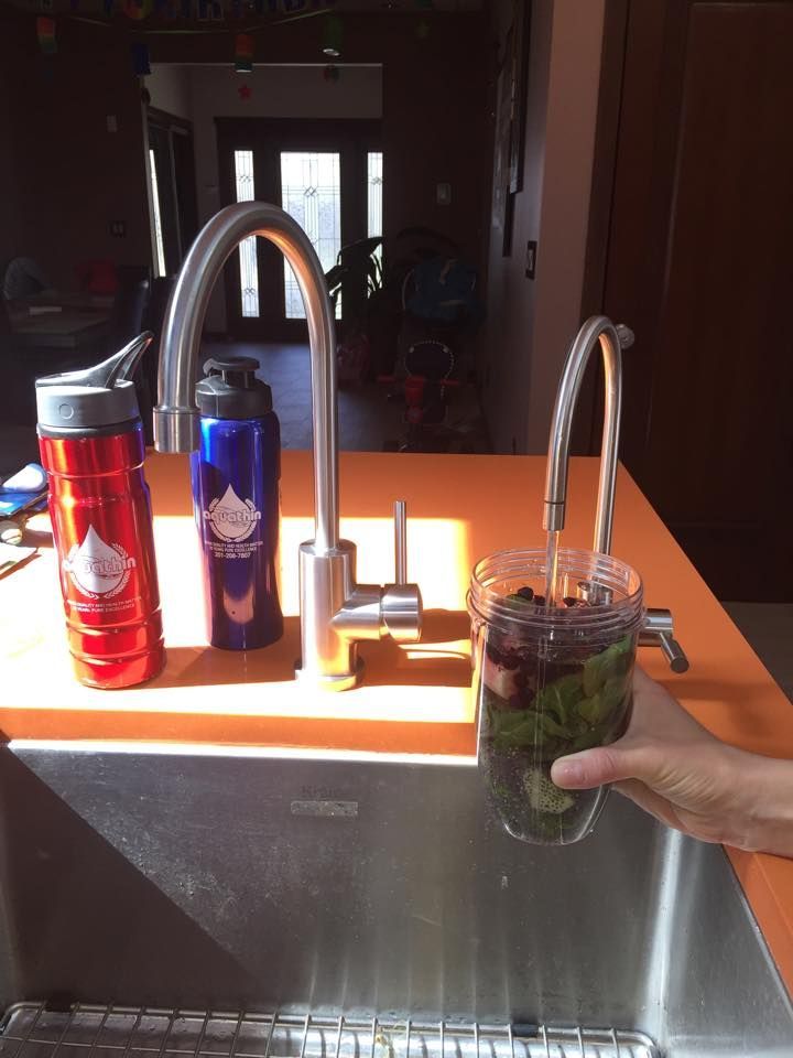 A person filling a glass of water with greens and berries from a kitchen faucet, next to two water bottles.