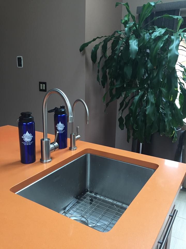 Stainless steel kitchen sink in orange countertop with two water bottles, faucet, and green plant.