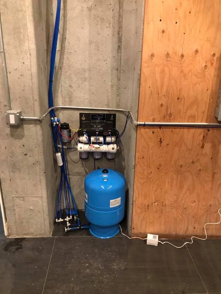 Water filtration system with blue tank, white filters, and pipes installed on concrete and wood walls.