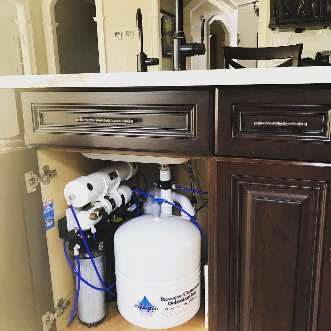 Undersink water filtration system with white tank, blue tubes, and black and white components under a dark wood cabinet.