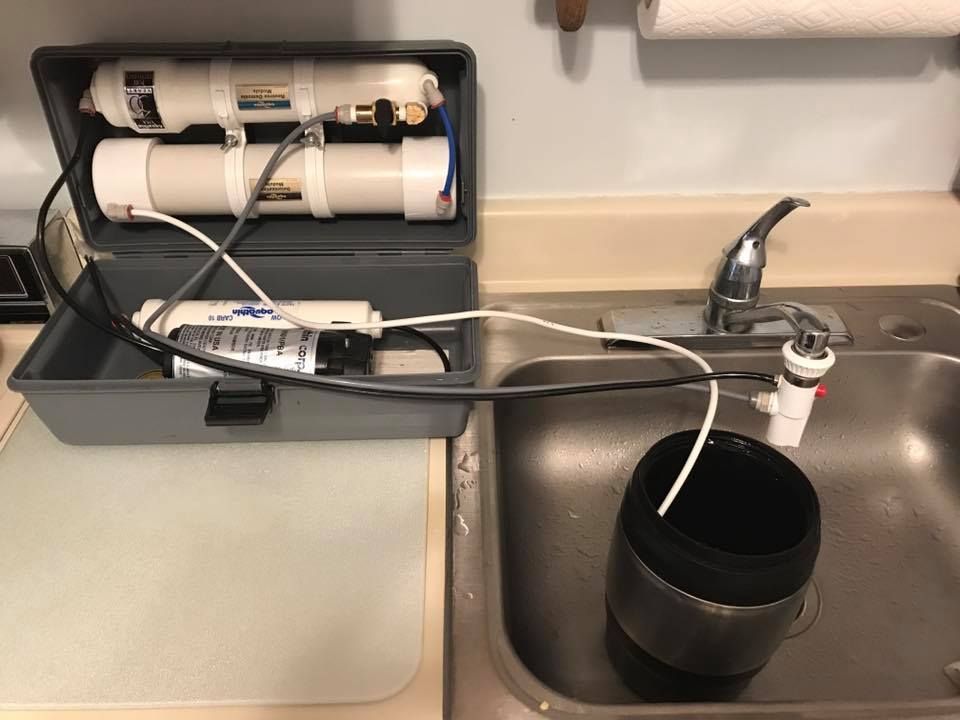 Water filtration system set up in a kitchen sink. Grey case holds filters connected to faucet and a bucket.
