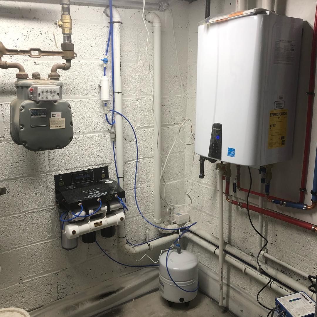Gas meter, water filter, and tankless water heater mounted on a white brick wall. Blue and copper pipes are visible.