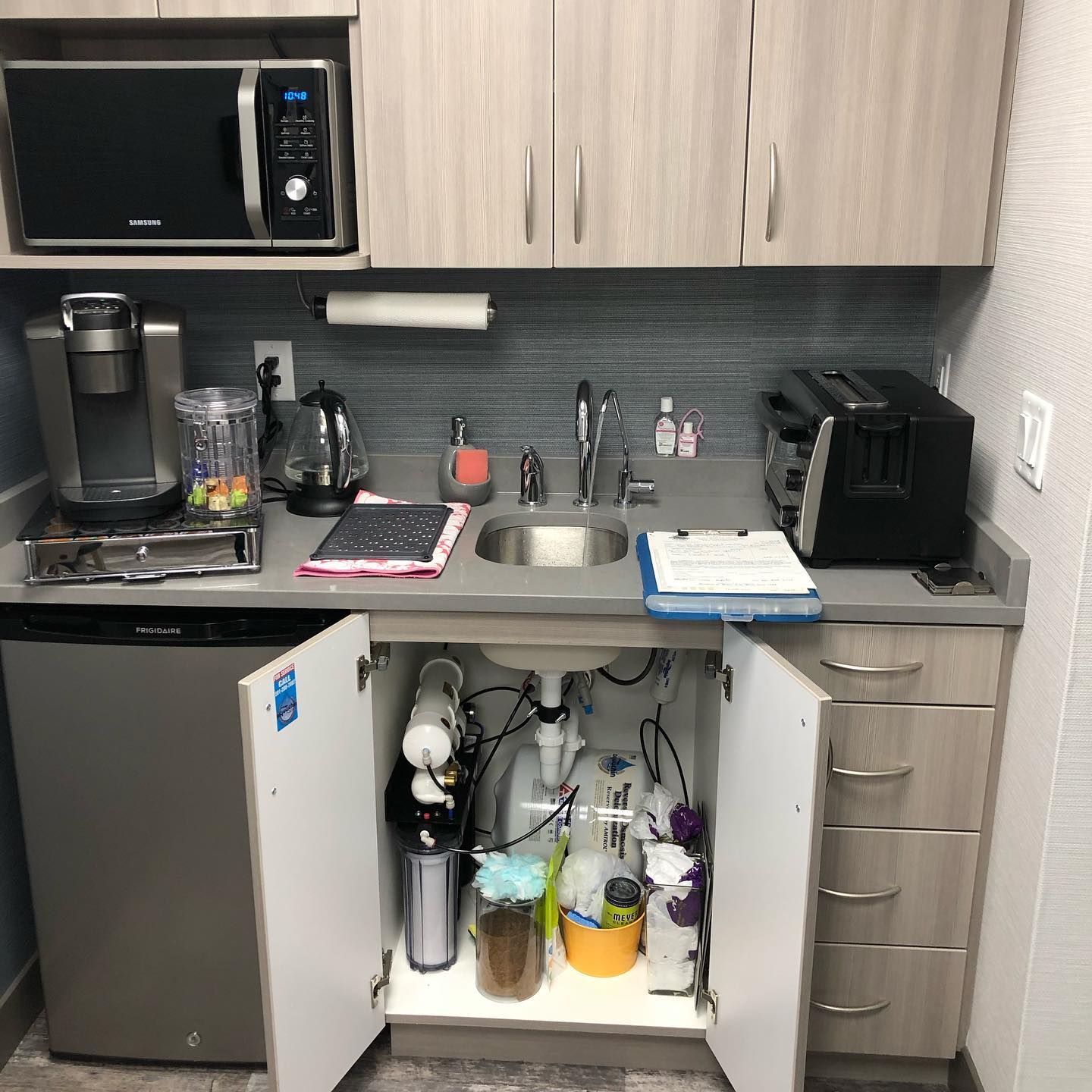 Small office kitchenette with microwave, sink, coffee maker, and mini-fridge. Cupboards are open underneath.