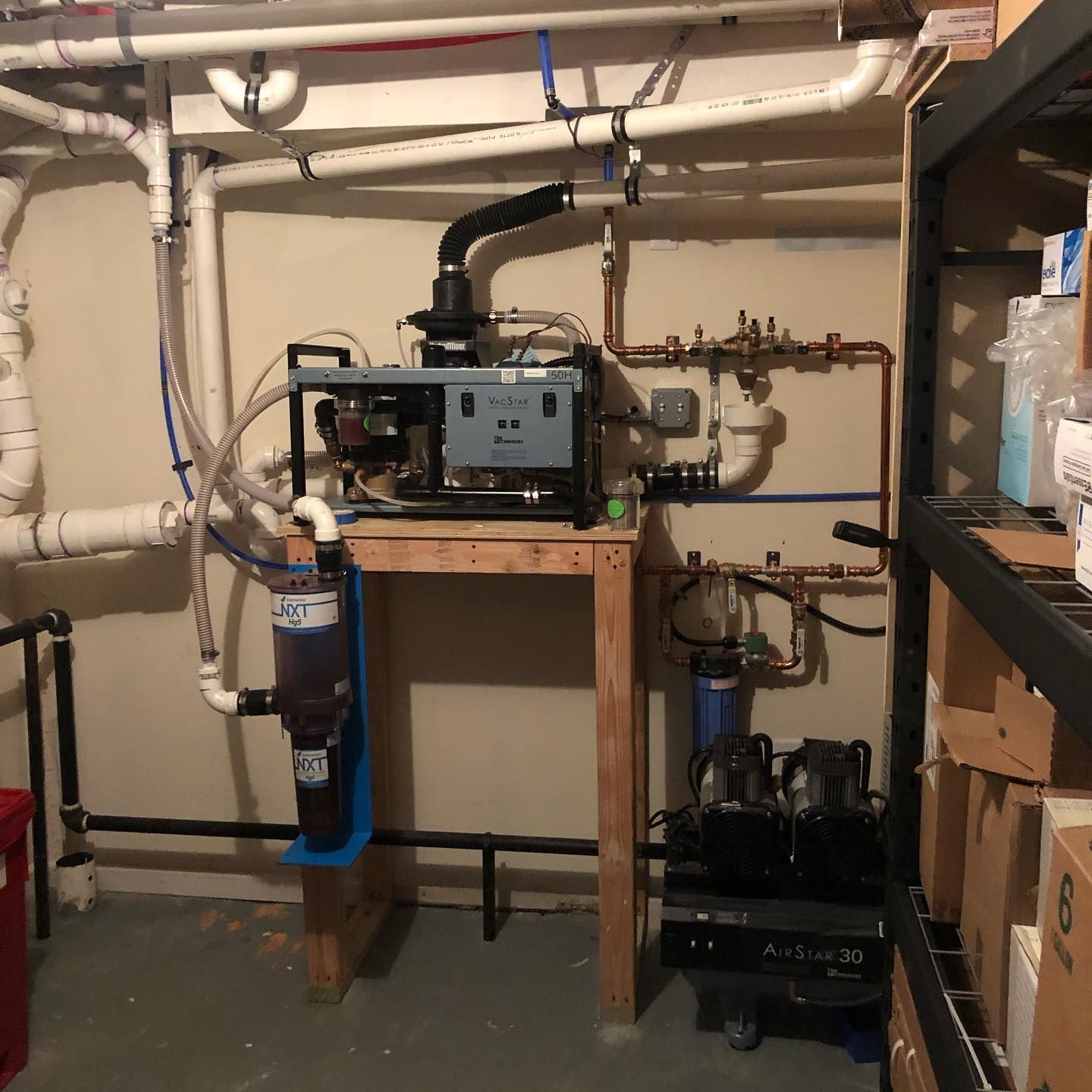 Basement with plumbing and water filtration system on a wooden stand near shelving.