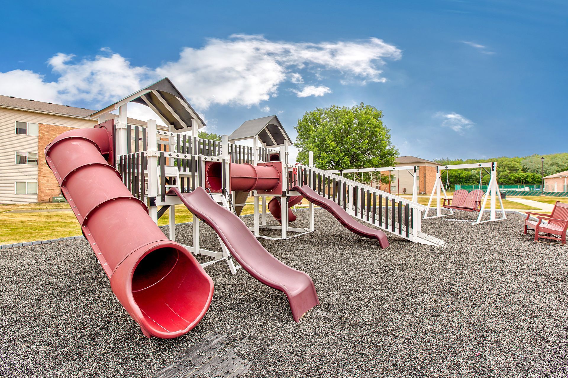 Playground with slides, tunnels, and swings; brown, white, and red structures on gray surface under blue sky.