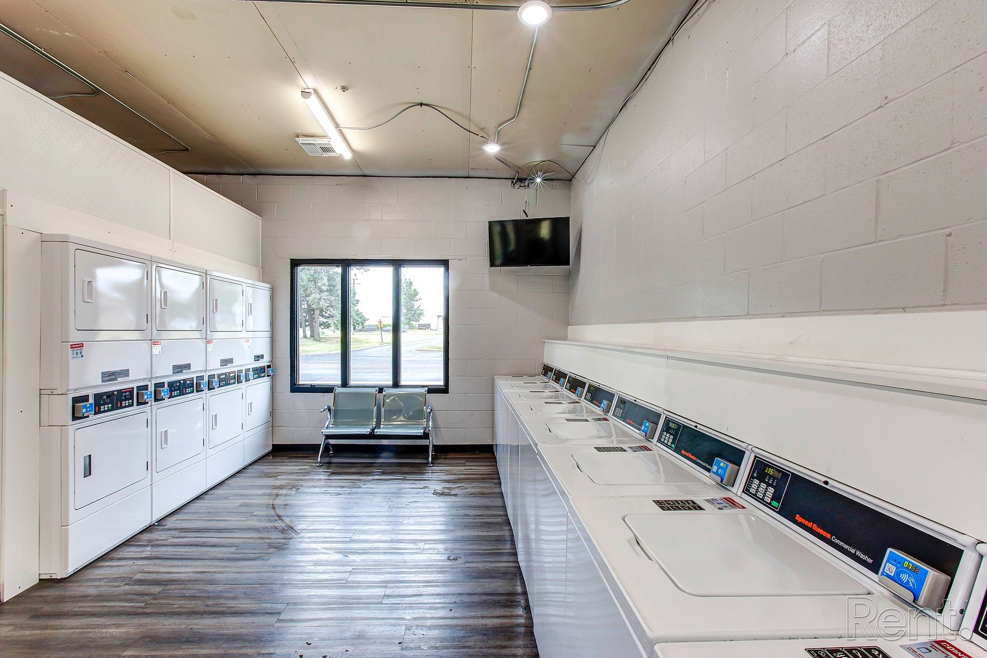 A laundromat with white washers and dryers, a window with chairs, and a TV on the wall.