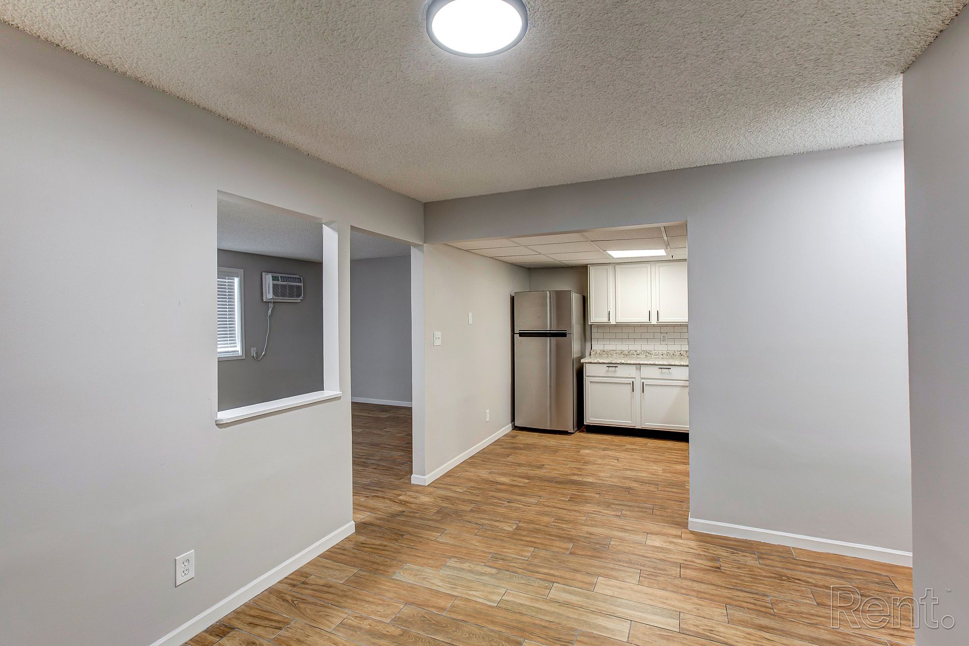 Interior view of a living space with light gray walls, a kitchen in the background, and cork flooring.