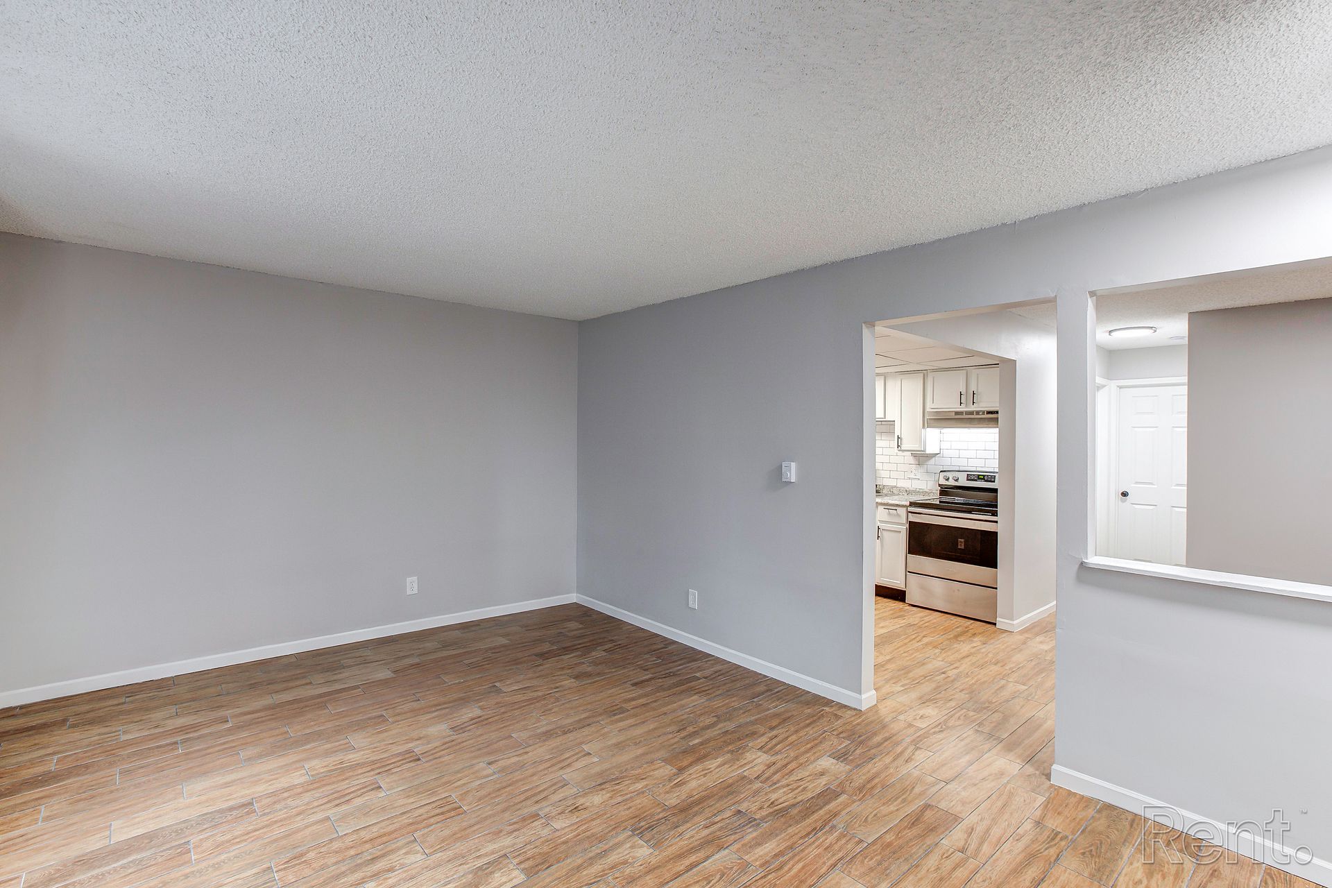Empty living room with light wood floors, gray walls, and a view into the kitchen.