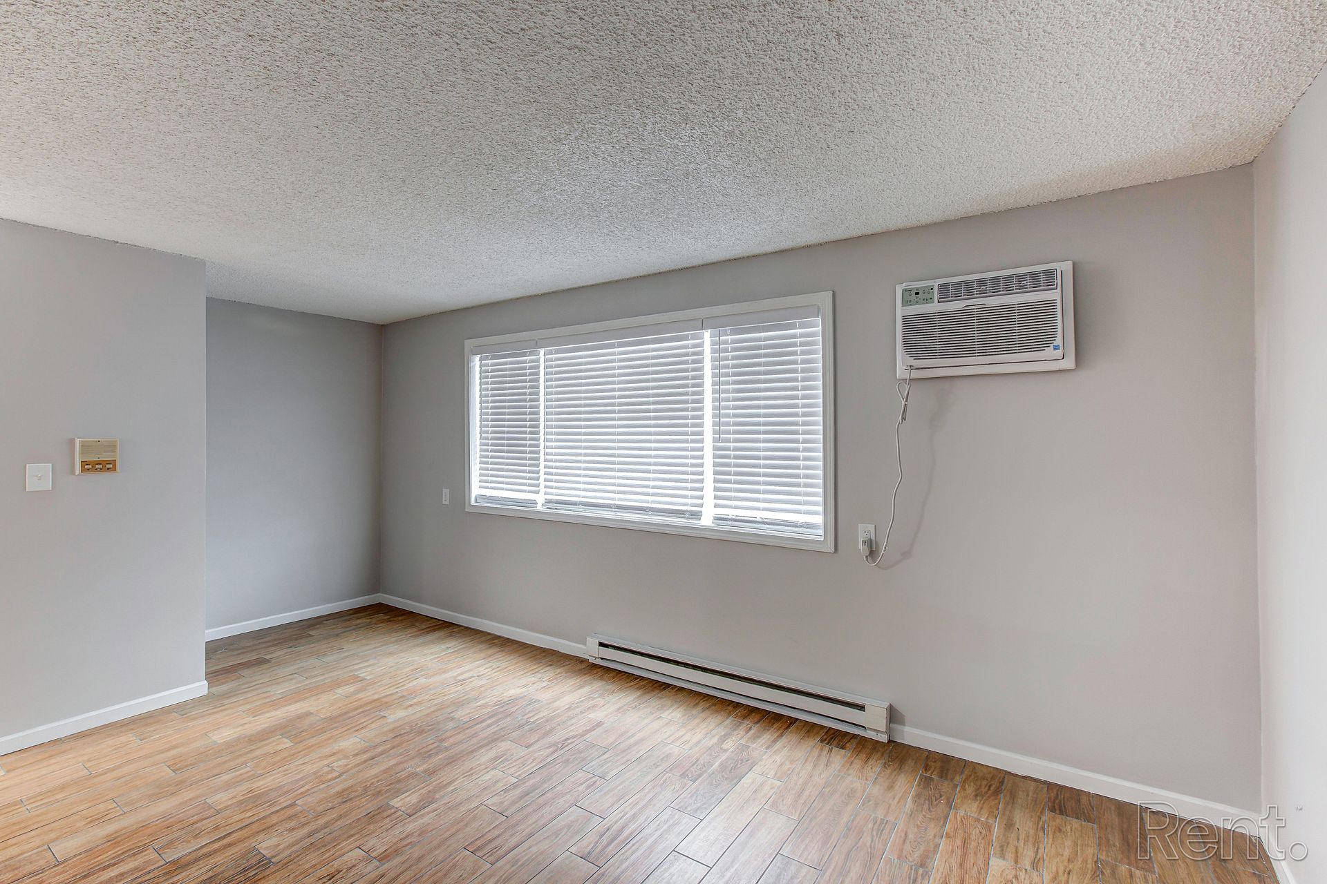 Empty room with wooden floor, a window with blinds, and an air conditioner. Gray walls and popcorn ceiling.