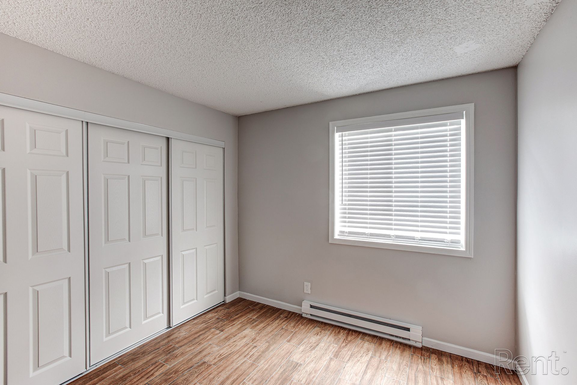 Empty bedroom with white sliding closet doors, a window with blinds, and wood-look flooring.