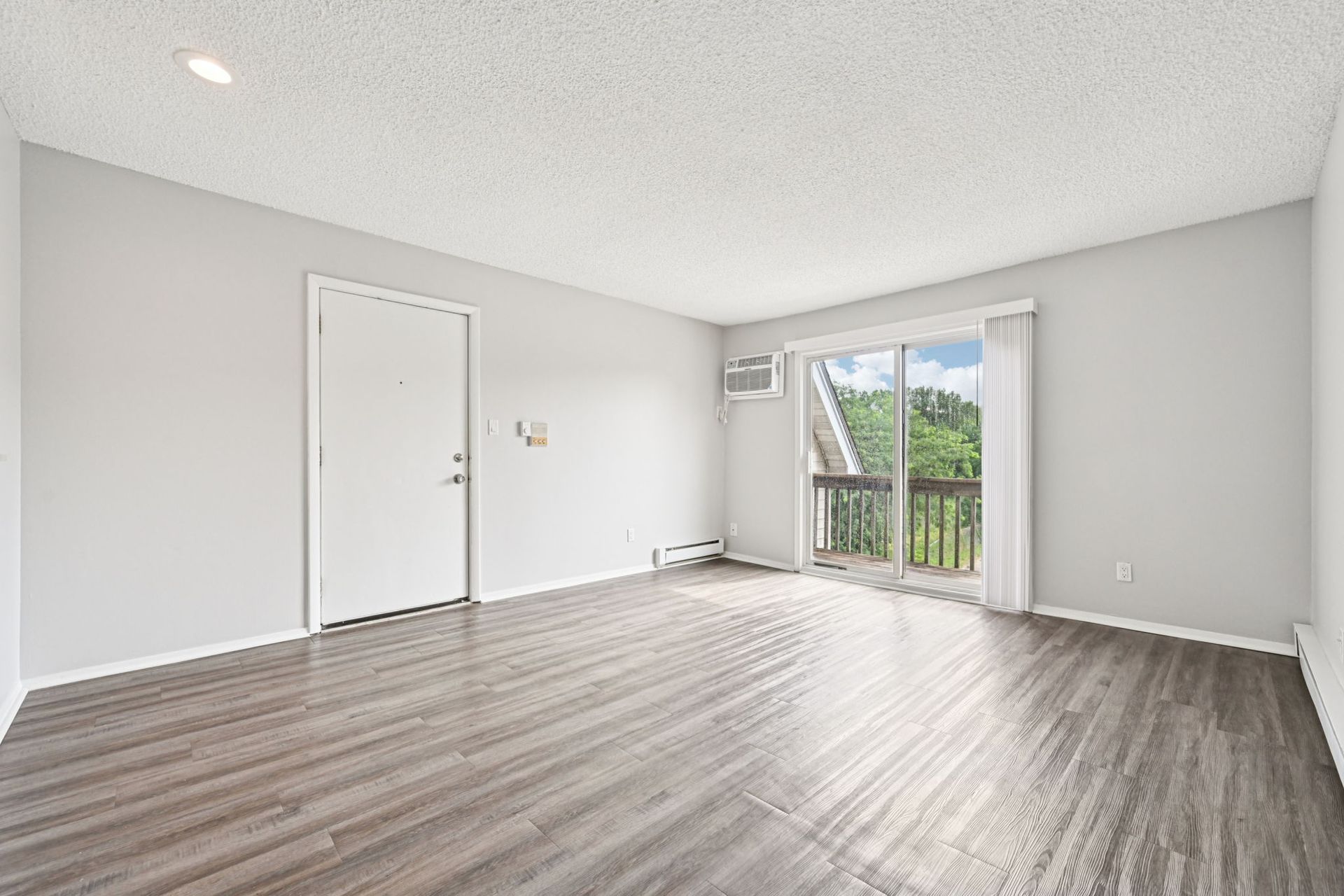 Empty living room with gray walls, wood floors, white door, and sliding glass door to balcony.